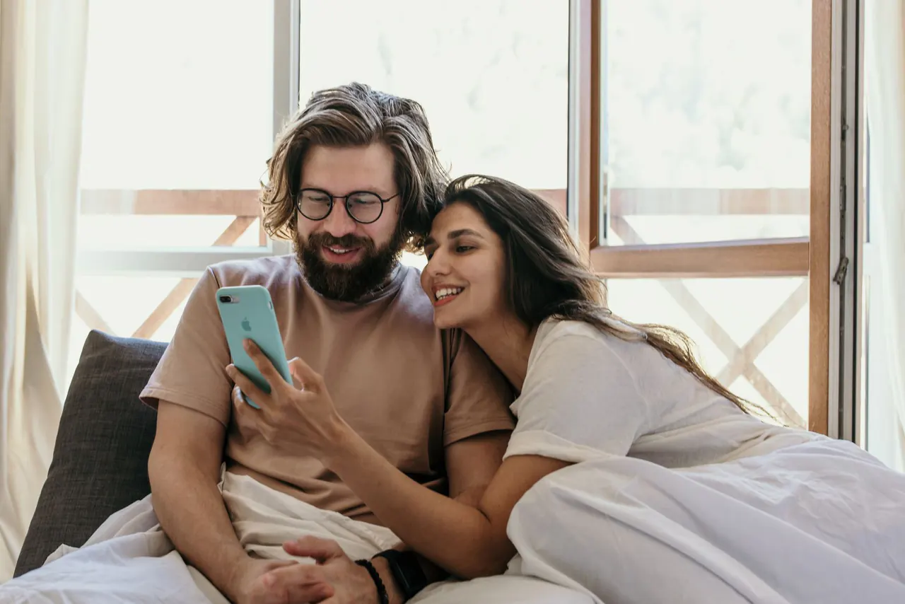 זוג מחייך מול הטלפון, יושבים ליד חלון ביום שמש